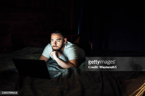 thoughtful man using laptop while lying on bed at home - dark room stock-fotos und bilder