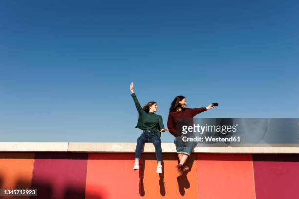 carefree female friends taking selfie while sitting on retaining wall - sitting on wall stock-fotos und bilder
