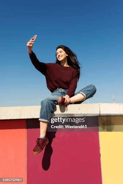 young woman taking selfie through smart phone on retaining wall - sitting on wall stock-fotos und bilder