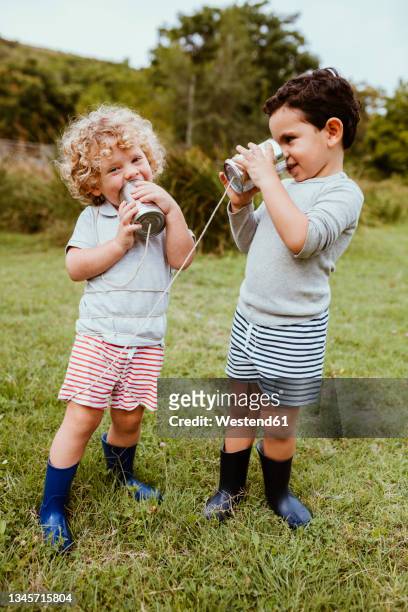 happy male friends talking through tin can phone while playing on meadow - tin can phone stock pictures, royalty-free photos & images