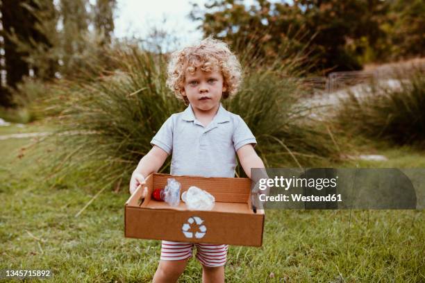 blond boy with curly hair carrying cardboard box with plastics on meadow - boy in a box stockfoto's en -beelden