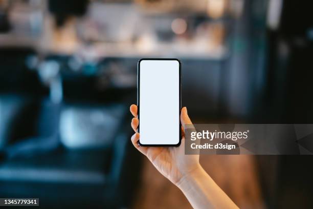cropped shot of a woman's hand holding up a smartphone with blank white screen in the living room at home. lifestyle and technology. smartphone with blank screen for design mockup - celular fotografías e imágenes de stock