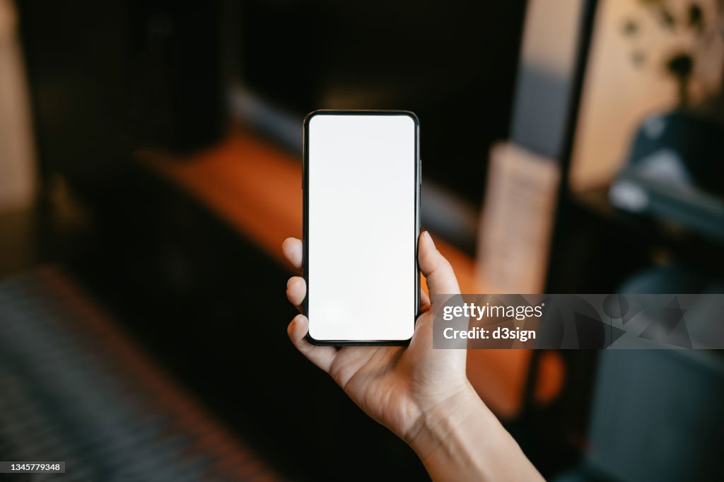 Cropped shot of a woman's hand holding up a smartphone with blank white screen in the living room at home. Lifestyle and technology. Smartphone with blank screen for design mockup