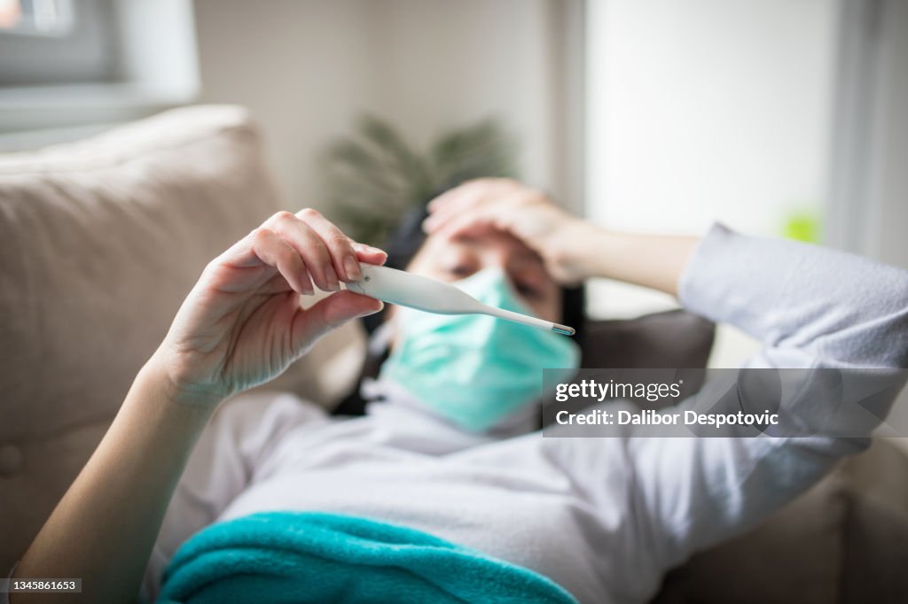 Young woman is looking at the thermometer. She has fever.