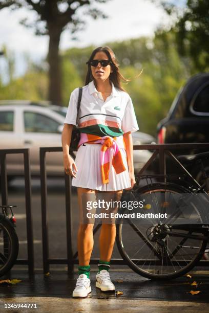 Guest wears sunglasses, a white with dark green and pink straps polo from Lacoste, a pale pink and orange striped wool pullover, a white pleated /...