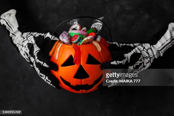 jack o' lantern, trick or treat bucket, filled with candies held by a skeleton hands on black background. halloween celebration concept. - black jack hand stock-fotos und bilder