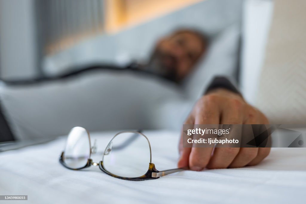 Overworked man lying on bed and holding his eyeglasses in hand.