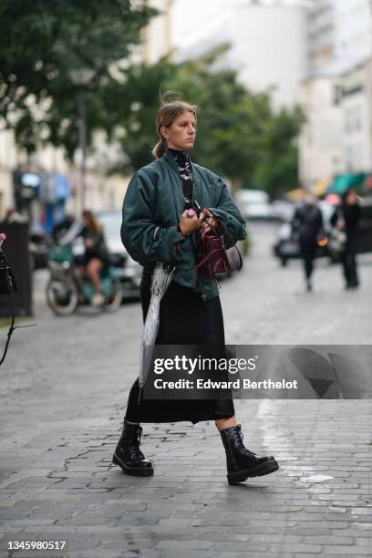 Guest wears silver earrings, a black with white print patter turtleneck pullover, a dark green bomber coat, a burgundy shiny leather handbag, a black...