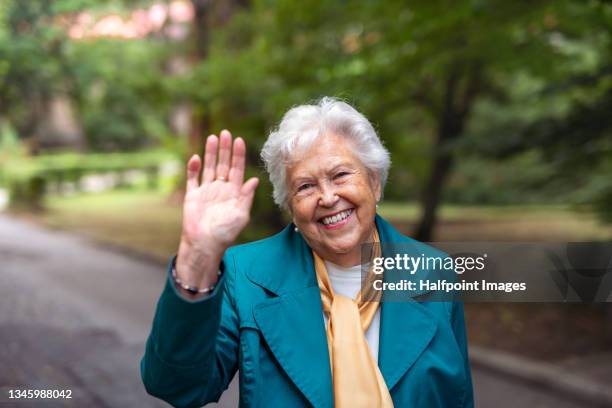 portrait of happy active senior woman on a walk in park, looking at camera and waving. - female waving on street stock pictures, royalty-free photos & images