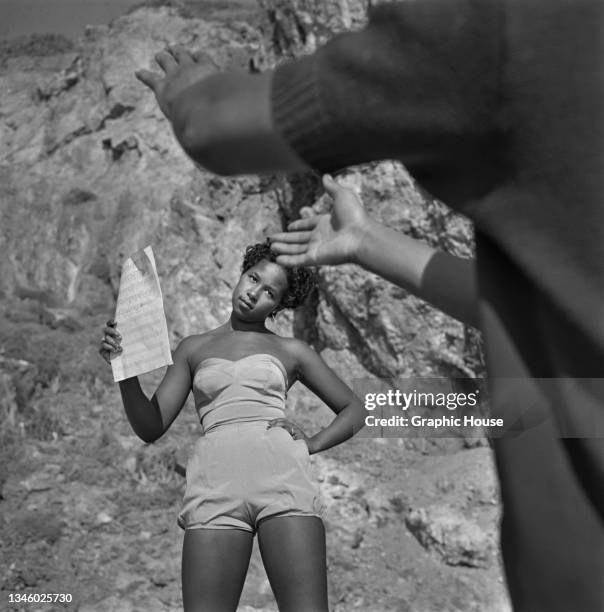 Young woman posing in a playsuit with a sheet of music for a photoshoot, circa 1950.