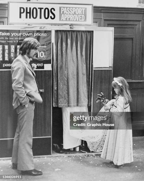 Bridesmaid holds the train for a bride who is having her photograph taken in a photo booth, USA, circa 1965.