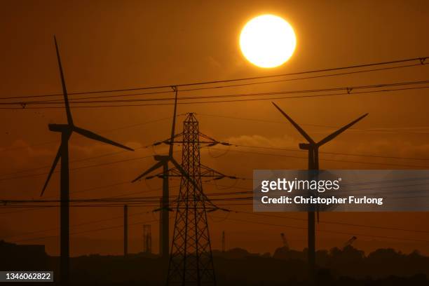 The sun sets behind wind turbines and electricity pylons dominate the landscape at Ince Salt Marshes near to chemical and manufacturing plants on the...