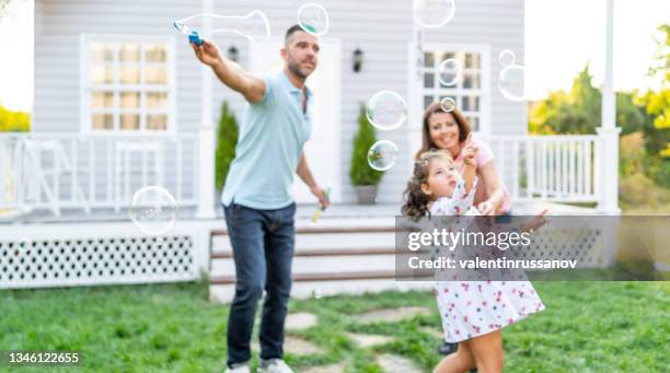 happy family playing soap bubbles on the grass in front of their porch. adorable little girl  playing soap bubbles with her mother and father outdoors. - bubbles happy stockfoto's en -beelden