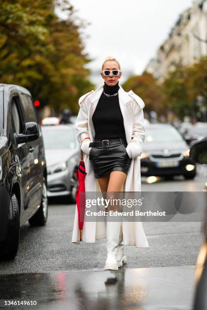 Elina Halimi wears white sunglasses, a silver large chain necklace, a black wool ribbed turtleneck pullover, a white long wool trench coat, a black...