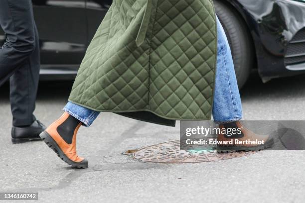 Guest wears a khaki quilted long coat, blue denim faded jeans pants, orange shiny leather ankle boots, outside Hermes, during Paris Fashion Week -...