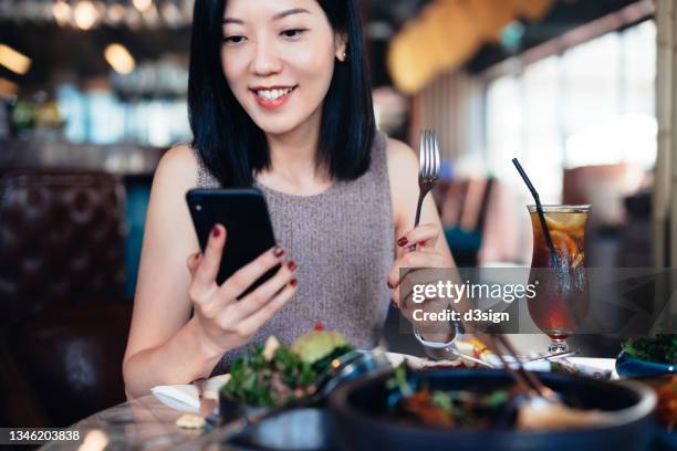 smiling young asian woman looking at her smartphone while sitting in restaurant with scrumptious meal freshly served on the dining table. eating out lifestyle. lifestyle and technology - paying for dinner imagens e fotografias de stock