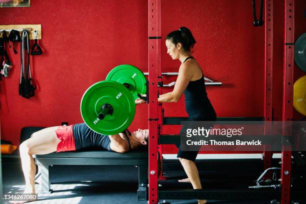wide shot of woman spotting friend bench pressing while working out in home gym - prop sporting position stock pictures, royalty-free photos & images