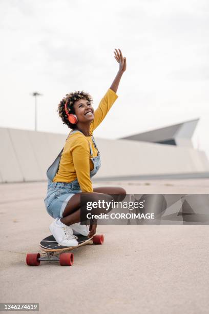 carefree afro skater woman enjoying skateboarding on the street while listening to music with headphones. - woman headphones sport smile iphone stock pictures, royalty-free photos & images