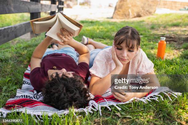young female same sex couple on picnic blanket with phone and book - sfogliare libro foto e immagini stock