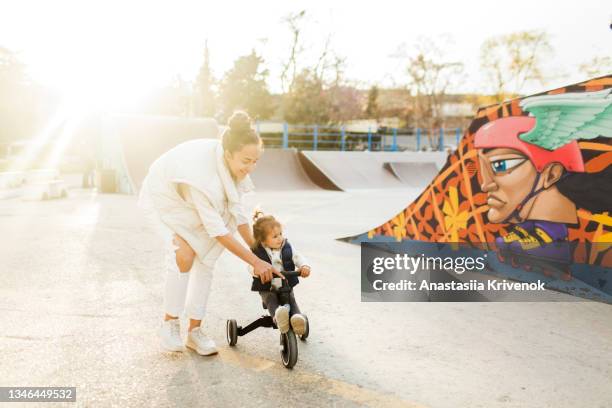 beautiful young mom teaching her little daughter how to ride a balance bike at the skate park in the city. - mother and daughter riding on skateboard in park stock pictures, royalty-free photos & images