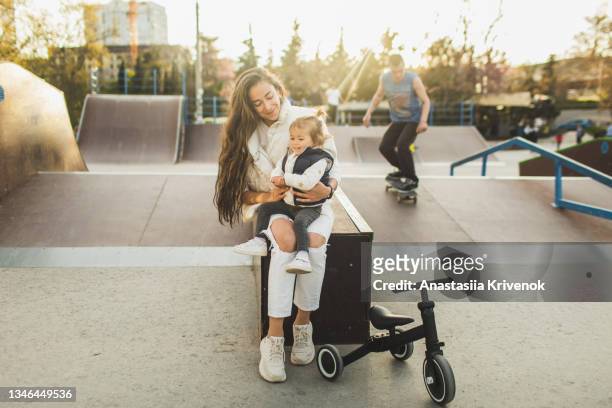 beautiful mother and daughter sitting at skate park together smiling happily - mother and daughter riding on skateboard in park stock pictures, royalty-free photos & images