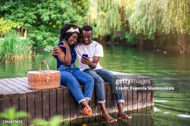 young black couple sitting on the pier in the park and looking photos in smart phone. - romantic picnic stockfoto's en -beelden
