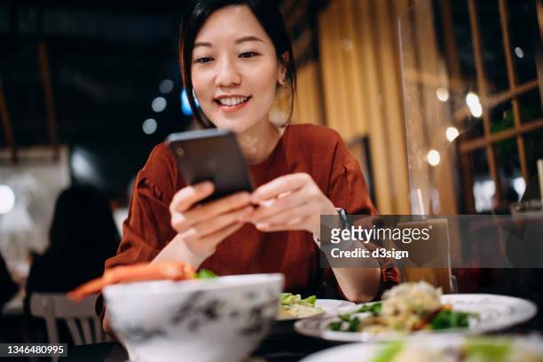 Beautiful smiling young Asian woman taking photos of scrumptious traditional Thai food served on dining table with smartphone while enjoying lunch in a Thai restaurant. Eating out lifestyle. Camera eats first culture