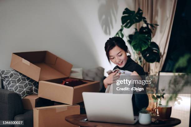young asian woman unboxing new purchase clothings from cardboard box that received from her online shopping retail delivery at home. she is happy and excited to see the content from the box. online shopping, trustworthy parcel delivery service - home shopping foto e immagini stock