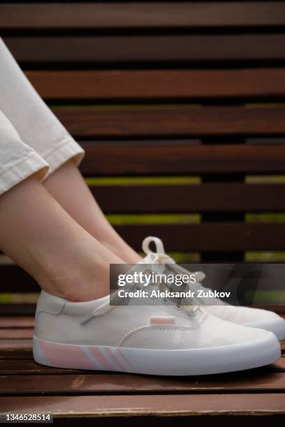 the legs of a teenage girl in new clean white sneakers on a wooden bench in a public park, on a summer day. a woman in modern fashionable shoes. youth culture. copy space. - female foot models fotografías e imágenes de stock