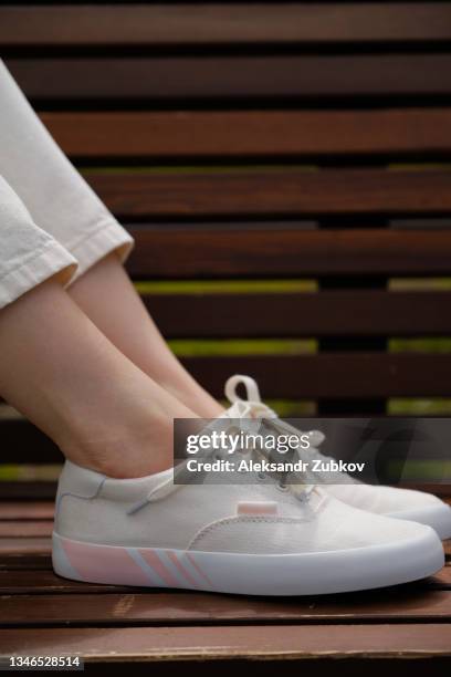 the legs of a teenage girl in new clean white sneakers on a wooden bench in a public park, on a summer day. a woman in modern fashionable shoes. youth culture. copy space. - female foot models ストックフォトと画像