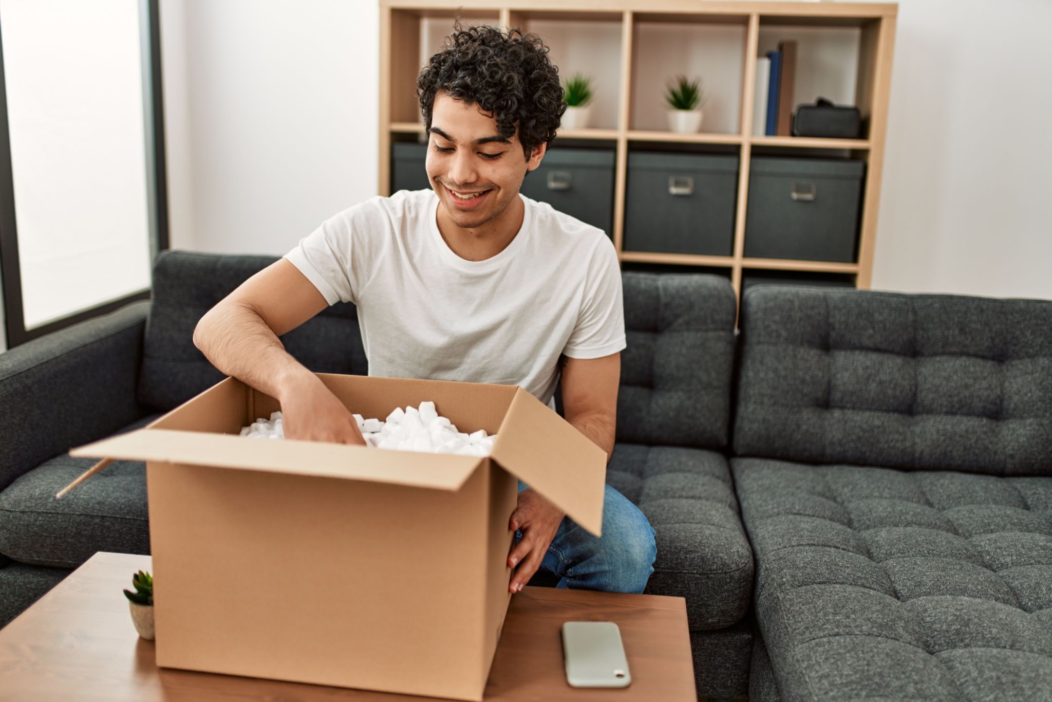 Young hispanic man unboxing cardboard box sitting on the sofa at home. Young hispanic man unboxing cardboard box sitting on the sofa at home.