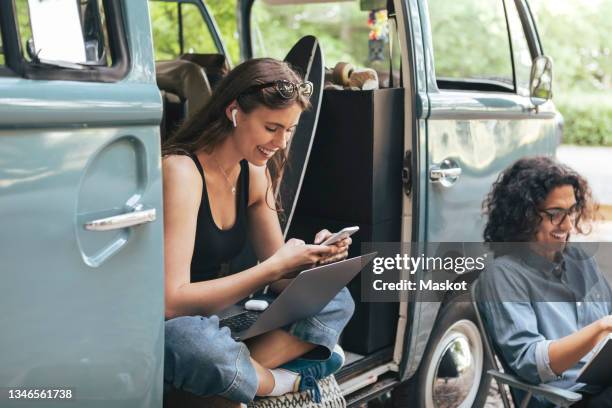 smiling woman using smart phone while male friend with book sitting by outside camping van - in ear headphones stock pictures, royalty-free photos & images