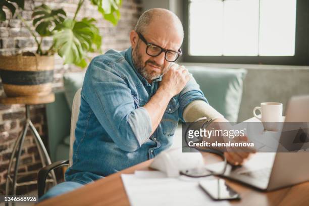 reifer erwachsener mann, der vom büro aus arbeitet (mit laptop, blutdruckmessgerät, überarbeitet) - hypertension stock-fotos und bilder