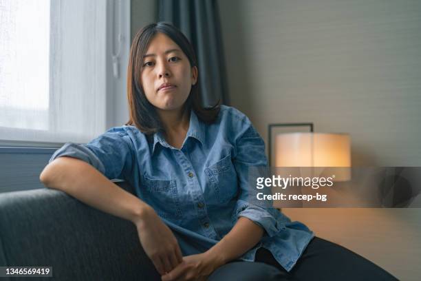 portrait of young woman sitting on sofa by window and looking at camera - stressed young woman sitting on couch stock pictures, royalty-free photos & images
