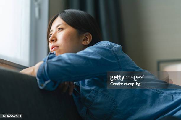young woman sitting on sofa by window and looking outside through window at home - expression stress stockfoto's en -beelden