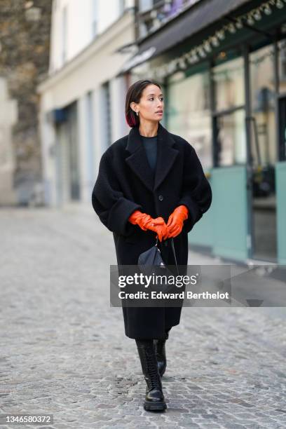 Alexandra Guerain wears silver earrings, a black t-shirt, a black wool oversized coat, neon orange shiny leather gloves from Prada, a black nylon...