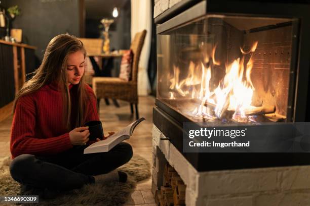 teenage girl enjoying a book and a cup of hot chocolate by the fireplace - sitting by fireplace stock pictures, royalty-free photos & images