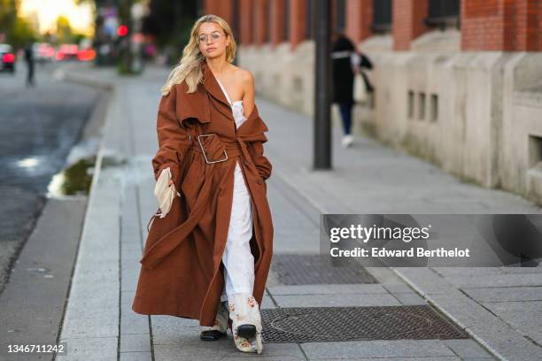 Stephanie Broek wears glasses, gold earrings, a white asymmetric shoulder shirt dress, a brown oversized long large belted coat, a beige with yellow...