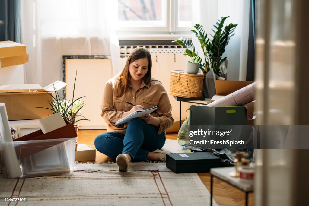 Ausziehen: Schöne lächelnde übergewichtige Frau, die auf dem Boden sitzt, umgeben von gepackten Kisten und eine To-Do-Liste macht