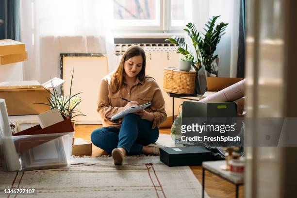 moving out: bella donna sorridente in sovrappeso seduta sul pavimento circondata da scatole piene e facendo una lista di cose da fare - trasloco casa foto e immagini stock