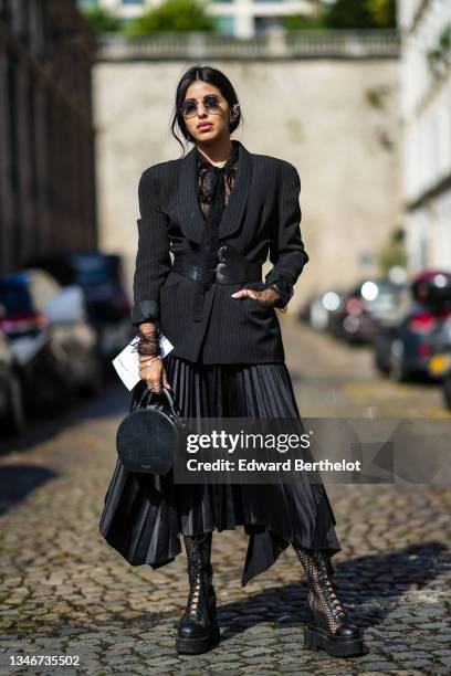 Guest wears black sunglasses, silver earrings, a black striped blazer jacket with shoulder-pads, a black lace pattern shirt with a knot neck, a black...