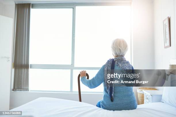 rückansicht einer nicht wiederzuerkennen älteren frau, die nachdenklich aussieht, während sie in ihrem zimmer im altersheim sitzt - rentnersiedlung stock-fotos und bilder