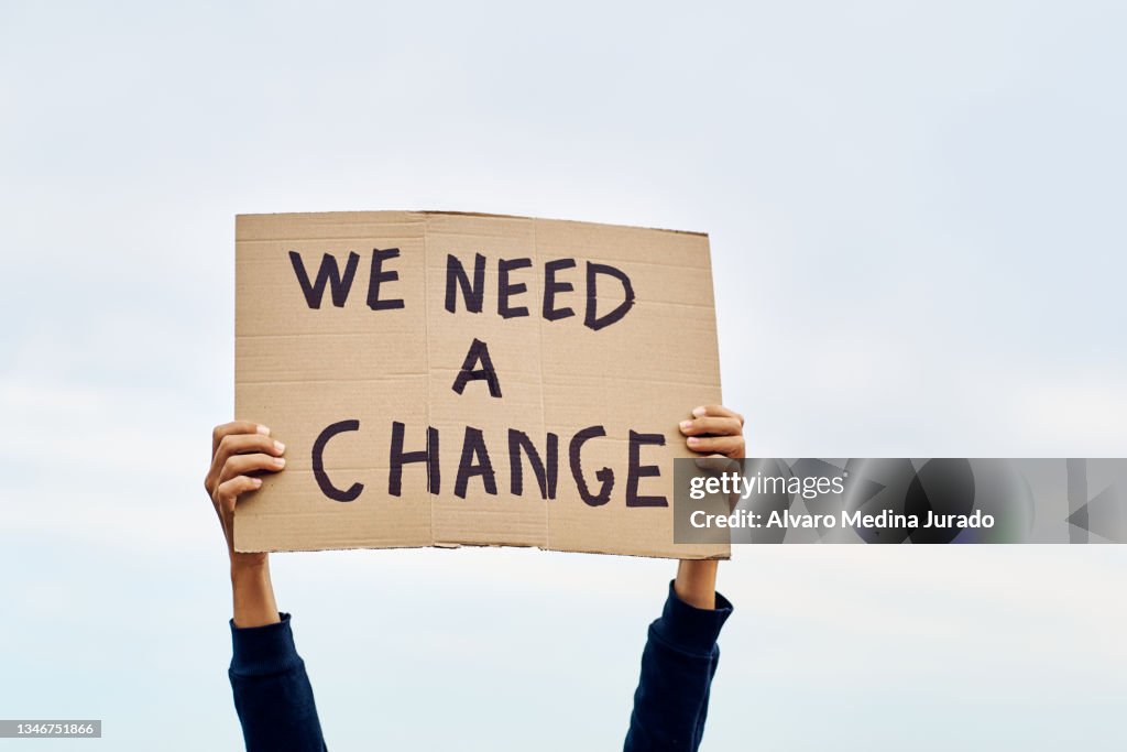 Unrecognizable woman's hands holding a protest banner with the message WE NEED A CHANGE, with the sky in the background.