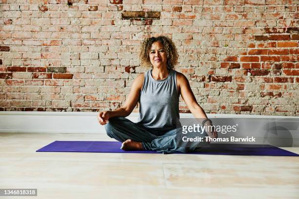 wide shot portrait of smiling mature yoga instructor seated in studio after class - schneidersitz stock-fotos und bilder