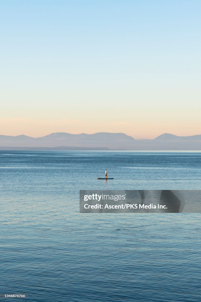 Man paddle boards through open water at sunrise