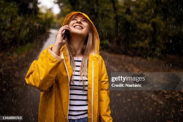 lächelnde junge frau im gelben regenmantel, die bei regen ein smartphone angreift und aufschaut - frau mit gelben regenmantel stock-fotos und bilder