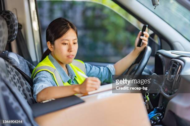 female delivery person sitting in van with parcels next to her and using smart phone to check and update orders - gig economy stock pictures, royalty-free photos & images