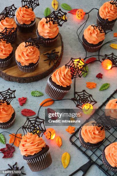 image of halloween chocolate cupcakes in paper cake cases on wooden cake stand and metal cooling rack, orange butter icing swirls topped with chocolate spider's webs, fondant autumnal leaves, plastic spider, string lights, elevated view - cupcake pattern stock pictures, royalty-free photos & images