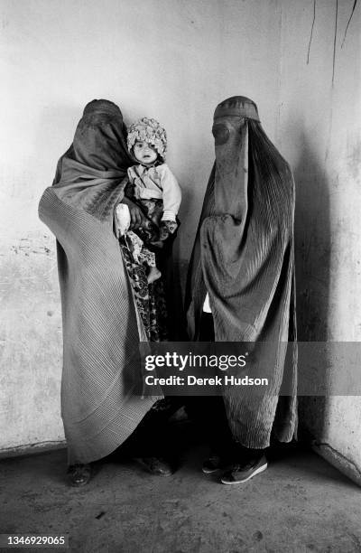 View of two women, both wearing burkas and one of whom holds a young girl in a bonnet, as they wait at a pediatric clinic, Kabul, Kabul Province,...