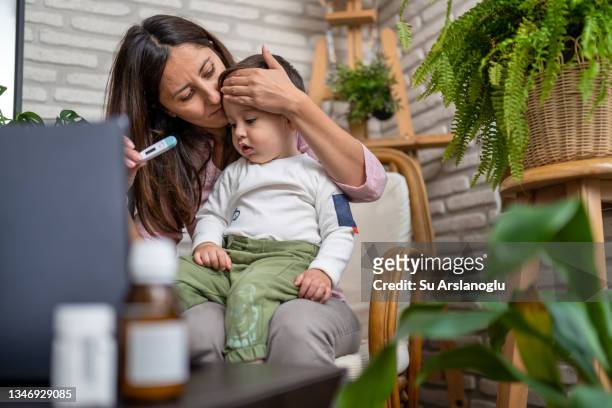 worried mother talking to doctor on laptop and taking baby's temperature during telemedicine session - woman home with sick children imagens e fotografias de stock